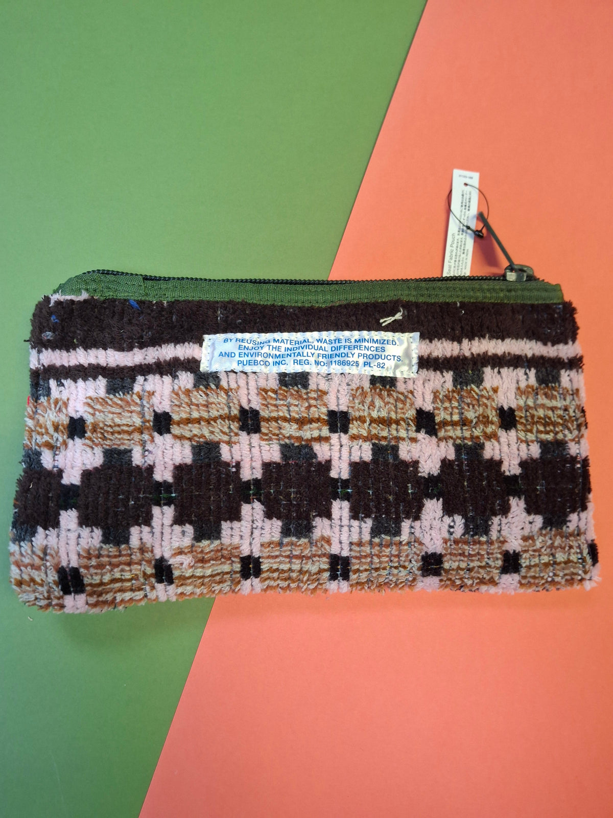A woven zippered clutch bag with a geometric pattern in brown, beige, and light pink, featuring a white brand label and a hanging tag, set against a diagonally split orange and green background.