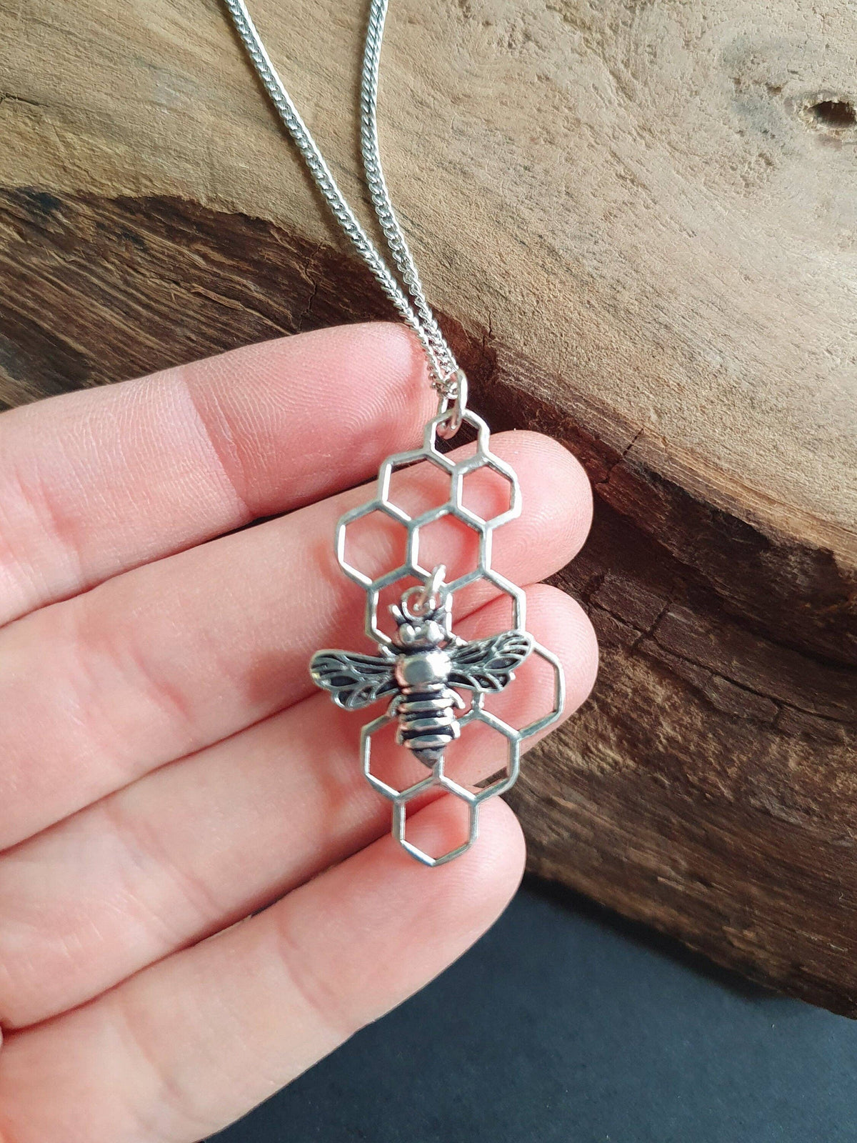 A silver-toned bee and hexagon pendant necklace held in a hand against a rustic wood background.