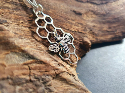 A silver-toned necklace featuring a honeycomb pattern and a small bee pendant, displayed on a textured piece of wood.
